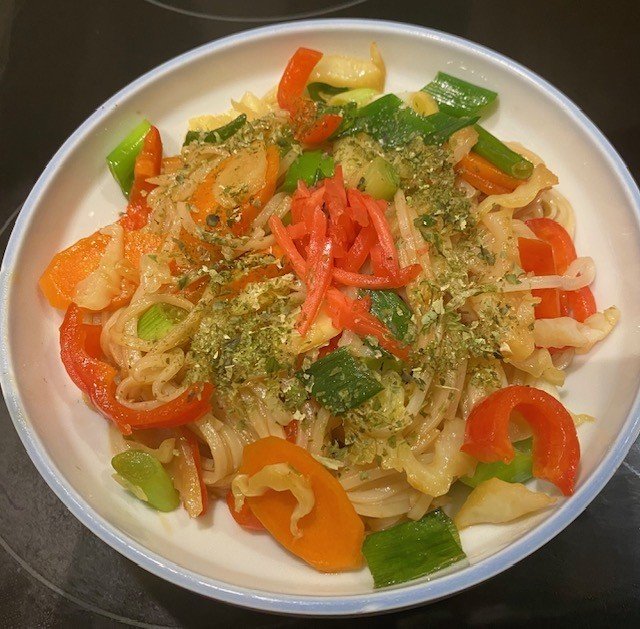 Gluteeniton yakisoba valkoisessa kulhossa: riisinuudeleita, oransseja porkkanaviipaleita, kaalia, vihreää purjoa ja punaista paprikaa, päällä punaista benishouga-inkivääripikkelssiä ja aonori-merilevähiutaleita